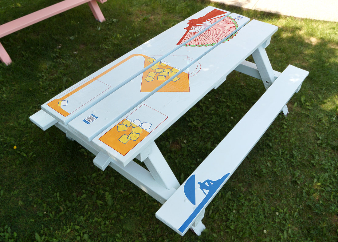 Picnic table painted with different designs. A pitcher of juice and glasses filled with ice, a hand holding a watermelon fan and someone reclining in the shade of an umbrella