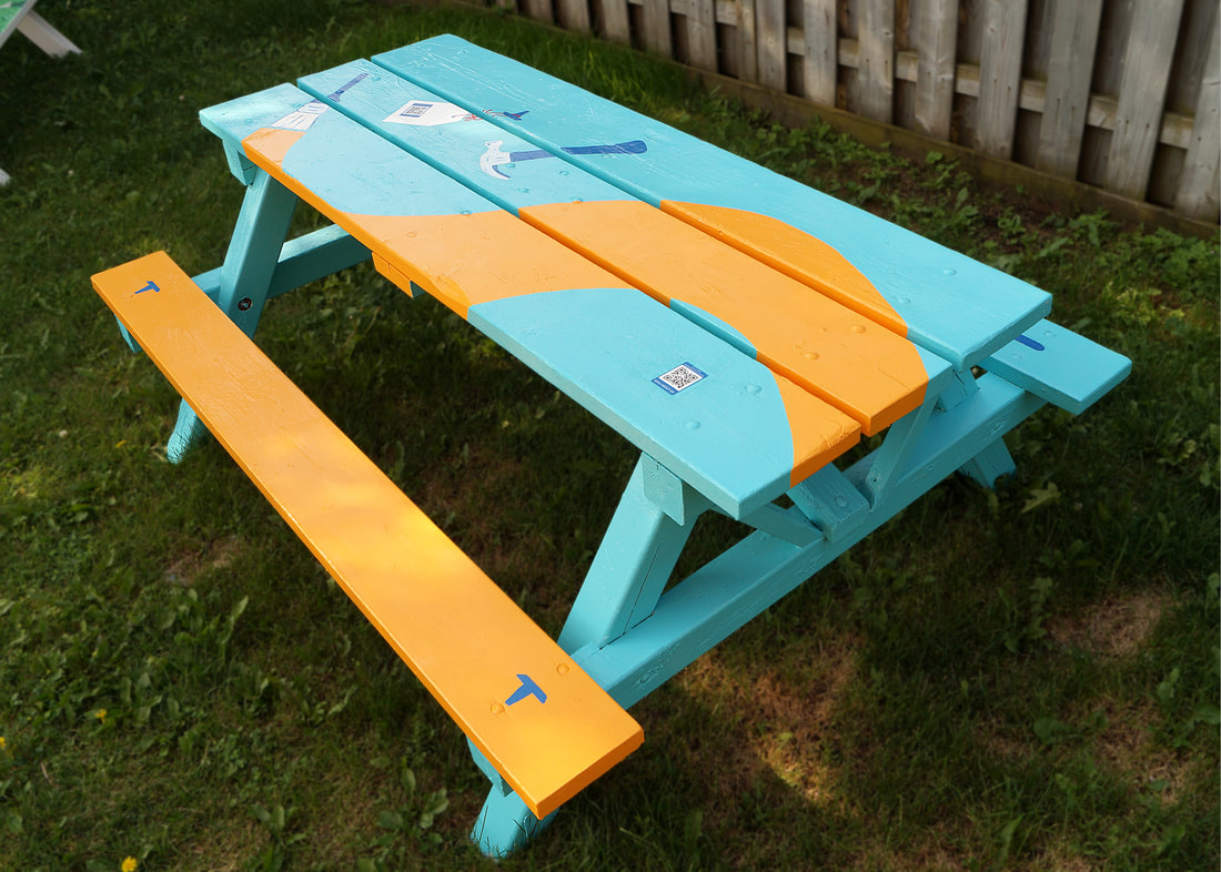 Picnic table painted to look like a paintbrush is covering it in orange paint. A hamer is poised to drive nails in